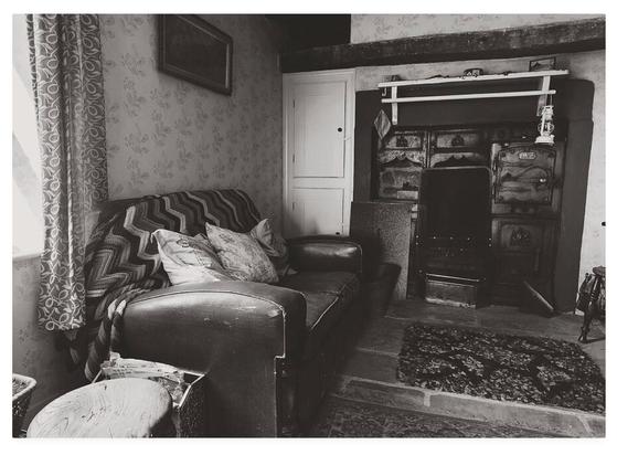 Black and white photograph showing a timeworn old parlour, with flagstoned floor and black cast iron range.  An old leather couch sits empty.