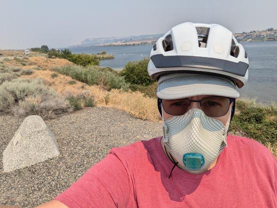 A person wearing sunglasses, a peaked cap, bike helmet and vented N100 mask in front of a river with a hazy sky behind.