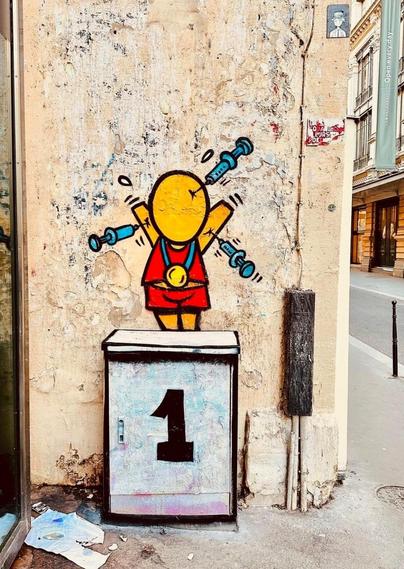 Streetartwall. On the corner of a street wall in Paris, a funny mural with a comic figure was sprayed for the Olympic Games. The figure (called Gouzou) is a small yellow man without a face, hands or feet. Wearing a red sports outfit, the Gouzou stands on a distribution box with a 