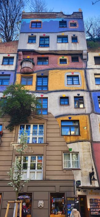 Colour photo of a very decorative and colourful building in Vienna, Austria. To the left is a tree and more trees grow on the roof garden. There are a variety of window shapes, designs and sizes. A person can be seen street level, it's a clear blue sky.