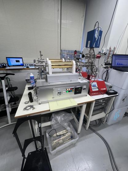 A laboratory where a rectangular machine sits on a table. The base of the machine is a little larger than a PC turned on its size and a gold coloured cylinder in mounted on top. Metal pipes lead off from the right. There are two laptops showing data.

