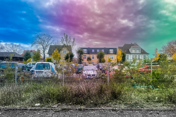 The image shows a suburban scene with several houses and a parking area filled with various cars. The sky above is cloudy and has been edited with a gradient of colors, transitioning from blue on the left to purple in the center and green on the right. In the foreground, there is a fence with some overgrown plants and grass. The houses are surrounded by trees with yellow autumn leaves.