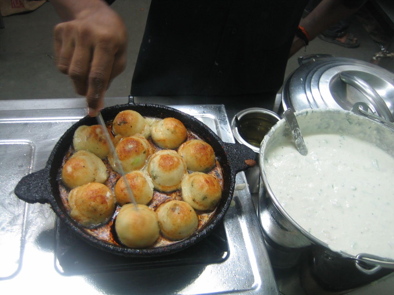 Kuzhi paniyarams being fried in a pan with semispherical depressions with a big bowl of white batter next to it