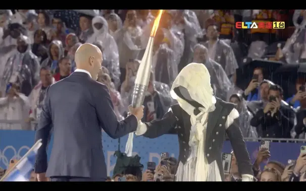 The masked torch runner passes the Olympic torch to Zinedine Zidane at the Paris 2024 Summer Olympics opening ceremony