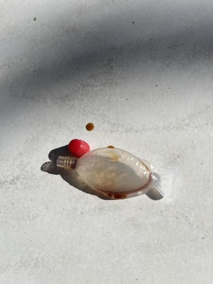 A small plastic fish-shaped soy sauce container with a red cap and a drop of soy sauce on a white surface.