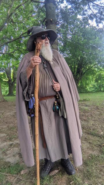 middle aged man with grey beard & sunglasses in Gandalf style outfit with staff standing in grove of trees