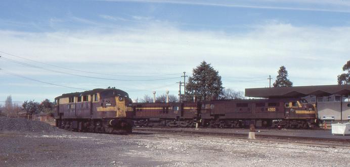 Three diesel locomotives, all in tuscan and yellow livery, sit in a railway yard with a 1960s style passenger railway station in the background.
