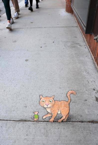 Streetart. A small cat and a mouse (named Eugene) were painted with chalk on a concrete sidewalk next to stores. The expansion joints were integrated into the picture. The orange and beige cat balances on the line next to the mouse in the green T-shirt. The mouse rests his arms on his hips and looks at the cat. Title: 