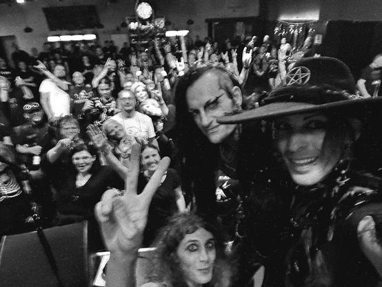 Black and white photo taken from the stage after the show with the band in the foreground and the audience in the background, everyone waving and smiling