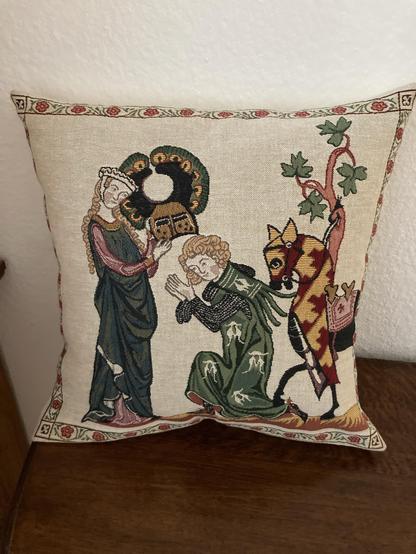 A decorative pillow with an embroidered medieval scene featuring a woman, a kneeling man, and a horse.