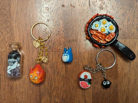 Tiny vial with different polished stones, keychains with painted clay to look like calcifer, soot gremlin, and a bento box. Tiny blue mini Totoro figurine and a black frying pan with bacon and eggs, with flames (calcifer) rising around the frying pan edges