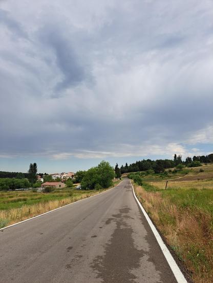 Fotografía de una carretera que pasa junto al pueblo de Griegos (Teruel, España), con algunas casas del pueblo visibles a la izquierda y el cielo nublado.