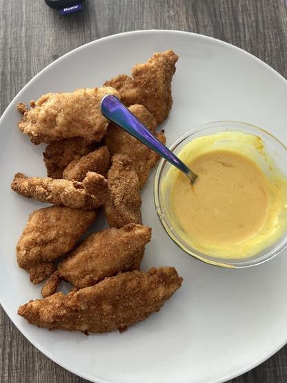 Crispy golden brown chicken strips with a bowl of yellow sauce on a white plate