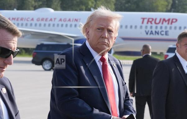The image shows Donald Trump standing on an airport tarmac, wearing a dark suit, white shirt, and red tie. He appears to be looking off to the side. His right ear is visible and has no markings at all that suggest any injuries. In the background, there is a plane with 