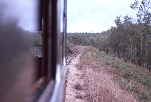 A view taken looking forward through an open window on the right hand side of a silver liveried Queensland Railways 2000 class railmotor about 3 windows back from the drivers cab.    To the left we can see back through the window in front of us and can just glimpse part of the cab and out through the front windscreen.  In front of the train we can see the lightly laid slightly curving narrow gauge track on which we are travelling.  At right is some low grass on a slope leading down to thickly wooded land in a gully.