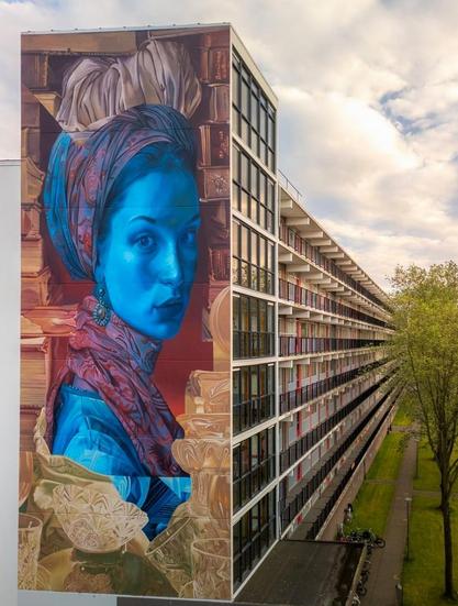 Streetartwall. The unusual mural of a young woman was sprayed/painted on the outside wall of a seven-storey modern building. In the style of old masters, a young woman is depicted looking at the viewer between walls full of books. The color of her skin is surprising. Her face is blue, as is her dress, which can be seen on her shoulders. Her hair is wrapped in elegant scarves and her neck is also covered by a patterned scarf in shades of red and blue. She is also wearing an elegant earring with a large round stone.  Her gaze is directed at the viewer. (The photo also shows the side view of the long skyscraper)
Info: Maria Tesselschade was a writer and artist from Alkmaar in the 16th century. She came from a family of writers with many talents and charisma. Unfortunately, there is no portrait of her, but she is said to have been a very attractive woman with charisma and wit. This mural is therefore a tribute to her.