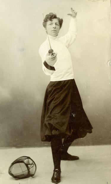 Janken Weil-Hansen in fencing outfit, pointing foil at camera, left arm & leg raised, fencing mask on floor 