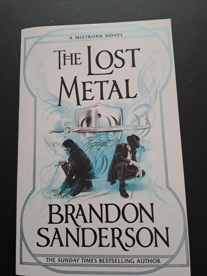 The cover of the book.

A man with a sword crouching on the left, a man with a bowler hat and walking cane crouching on the right. Behind them blue mist swirls in front of a sword and shield. The shield has Harmony's allomantic symbol on it.