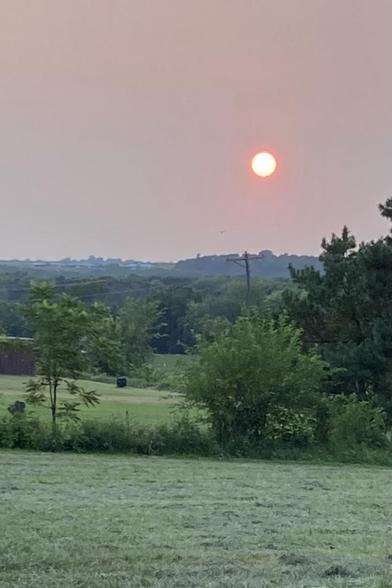 A red orange setting sun seen through haze of smoke from the Canadian wildfires.  Hills a couple of miles away are smoky blue.  Freshly cut hay in the foreground. 