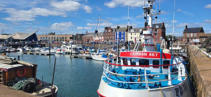 Colour photo of a fishing harbour in Arbroath,  Scotland,  taken today. 
Photo shows several fishing boats dockside with buildings in the background. It's a superb summer day, with cotton wool clouds around.