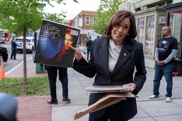 Kamala Harris in a suit smiles while holding an Alison Moyet vinyl record. She stands on a sidewalk with some people and buildings in the background.