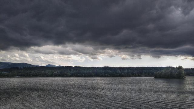 Photo of a lake, that water surface is perturbed by wind. There is a small is7alnd completely covered by trees. There are dark heavy clouds in the sky overhead, except far in the distance where it is almost clear.