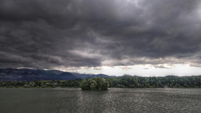 Photo of a lake with a small overgrown island and a mountain range in the background. There a dramatic thick dark clouds in the sky.