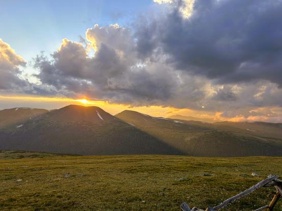 A photo of a sunset. The sun is just about to set at the crest of a mountain. Sunrays filter from the sides of the slopes on either side. Green grass sits in the foreground. The sky is partially clouded, but also a bit of blue showing through. 