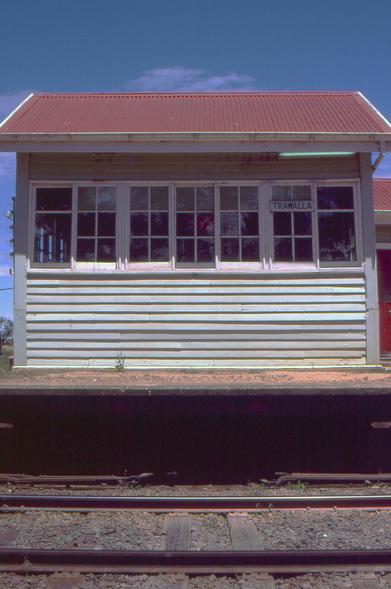 A front-on view of a weatherboard signal box standing on an earthen-topped passenger railway platform.  The signalbox, which is slightly elevated, has a lower part of horizontal light green coloured weatherboards, with  a fully glazed set of white painted wooden framed windows and a peaked red painted corrugated iron roof.  An enamel station nameplate with black letters on white reading TRAWALLA is affixed to the beading on one of the windows.  In the foreground running left to right is the main line track  We can see the gap beneath the platform through which the mechanical rodding and wiring accesses the signal box.