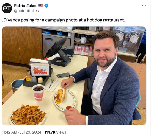 JD Vance trying not to look weird and failing...with a hot dog on a plate by the checkout register as if he's sitting at a table.