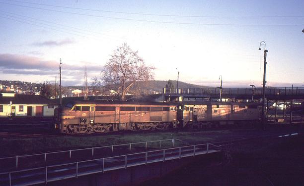 Looking across a railway yard, two carbody style diesel locomotives in Indian red livery with yellow lining sit coupled with the warm coloured morning sunlight illuminating this side.  The left loco has a streamlined cab facing left, whilst the right loco has flat cabs at both ends.  In the shade in the lower foreground is a turntable, and to the right, a footbridge crosses the yard.  A tree stands behind the locos and a hill and  the buildings of the city fill the background beneath a light blue, partly clouded early morning sky.