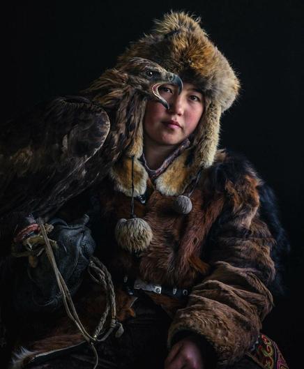 Photography. A color photo of a 14-year-old girl (named Aymoldir Daiynbek) in traditional Mongolian dress together with an eagle. The background is black. She is wearing a fur hat with earflaps and a fur coat with patterns in earth tones ranging from brown to beige. A majestic golden eagle sits on her left hand and its head covers her right eye. The bird has its mouth wide open as if it is screaming, an impressive portrait photo, perfectly staged. 
Info: Aymoldir Daiynbek is a 14-year-old eagle hunter from the remote Altai Mountains in western Mongolia. She has followed in the footsteps of her father and grandfather and was crowned winner of the Golden Eagle Festival in 2023, where hunters and their golden eagles are tested for speed, agility and accuracy.