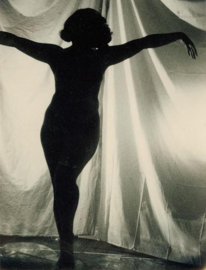 Black and white photo of a standing woman's black silhouette with arms splayed out on either side like she's about to take a bow, with white gauzy curtains behind her