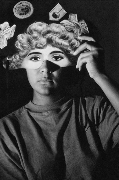 Black and white photo of a young Black woman in a baggy tshirt holding a half-mask over her eyes and forehead, decorated with wavy light hair and illustrated decorations