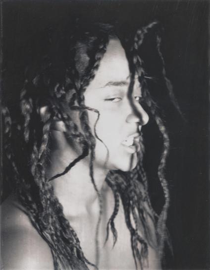 Black and white photo of a young Black woman with long braids falling askew over her face and shoulders. She looks sideways at the viewer with half-closed eyes and bared teeth