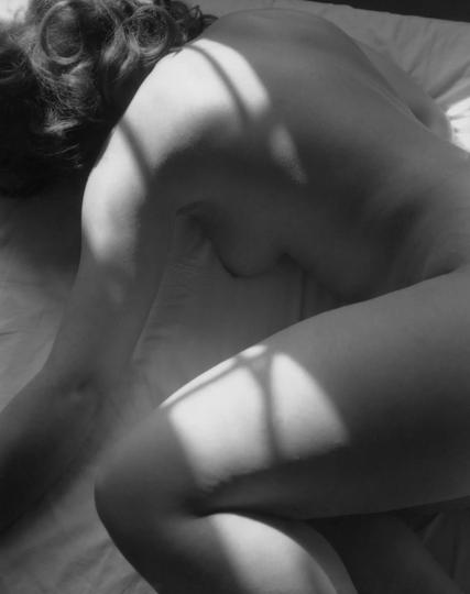 Black and white photo of a young Black woman lying nude on her stomach on a bed with white sheets, her legs curled up and her face hidden under dark wavy hair