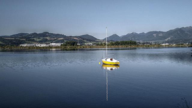 Photo of yellow-white sailboat on a lake. There is a mountain range and some smaller hills in the background. The sky is clear.
