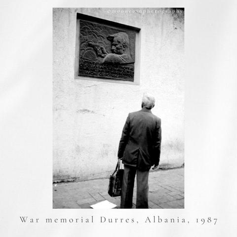 Older man with shopping bag in his left. hand, back towards the camera looks up at a WW2 Memorial with a man and gun inscription below.