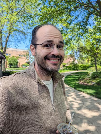 Steve sitting in a park with an iced cold brew coffee, wearing a quarter zip brownish gray sweater over a pale yellow tee. He's smiling directly at the camera with a hint of uncertainty. Lots of green in the background of both trees and grass, and a winding sidewalk through it all.