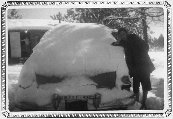 Young woman leaning on the side of a snow-covered Mercury car. In the background is a man standing in an open house door with 10 inches of snow on the roof.
