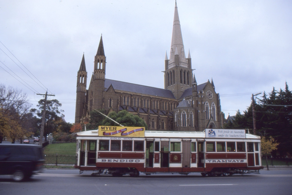On a dull day we see a brown and cream liveried dropcentre bogie tram with a  penny farthing wheelset beneath each end saloon and four openings in the dropcentre section runs left to right along a centre of road track with the leftmost trolley pole raised to the overhead wire.  In the background on a hill directly behind the tram is a large gothic style cathedral of mid grey coloured stone with a main spire of notably lighter coloured stone betraying it as a relatively newer addition. 