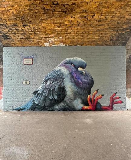 Streetartwall. The funny mural of a fat pigeon was sprayed/painted under an old stone bridge. The background is light gray. The fat pigeon is sitting on the ground, red feet forward and seems to be resting.