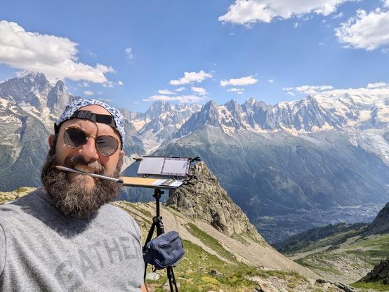 A photo of the mont blanc mountain range from across the valley, with myself and a painting sketch easel in the foreground. I've got a colored pencil in my mouth, a beard, and sunglasses on.