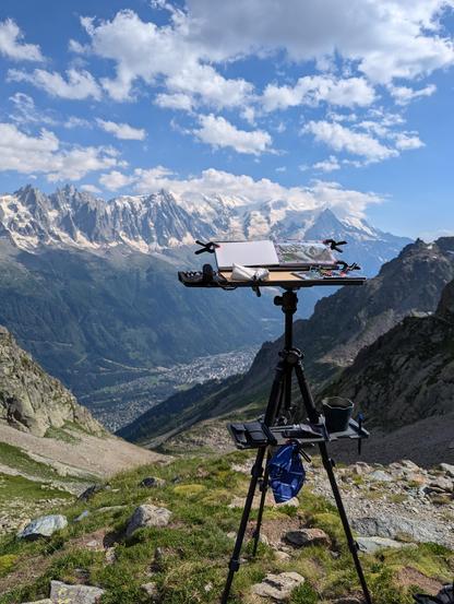 A photo of the mont blanc mountain range from across the valley, with a painting sketch easel in the foreground. The mountains are called with snow