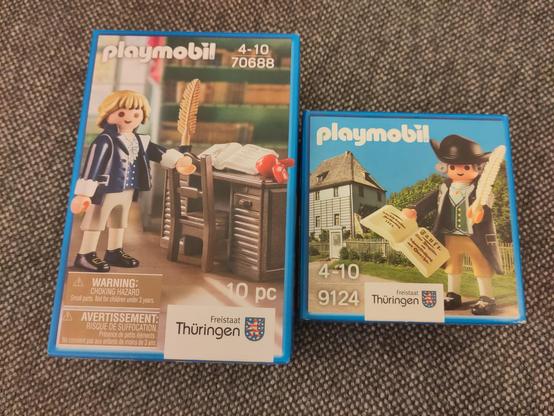 Two boxes of playmobil figurines, one depicts Friedrich Schiller at a writing desk, with a feather pen and a red apple, writing Wilhelm Tell. the other figurine is of J.W. Goethe, with a dashing hat and also a feather pen, he is proudly, some would say smugly, holding a copy of Faust. His garden house in Weimar is in the background