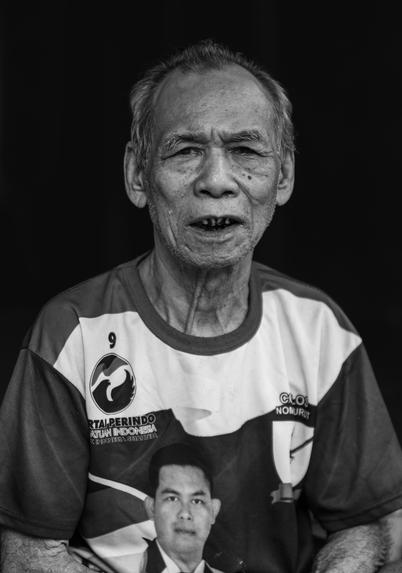 An elderly man wears a shirt with printed images and logos. The photo is in black and white.