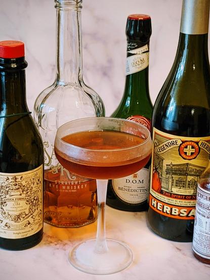 Cocktail in a coupe surrounded by bottles of ingredients - Sazerac rye, carpano vermouth, d.o.m Benedictine, herbsaint, Peychauds bitters