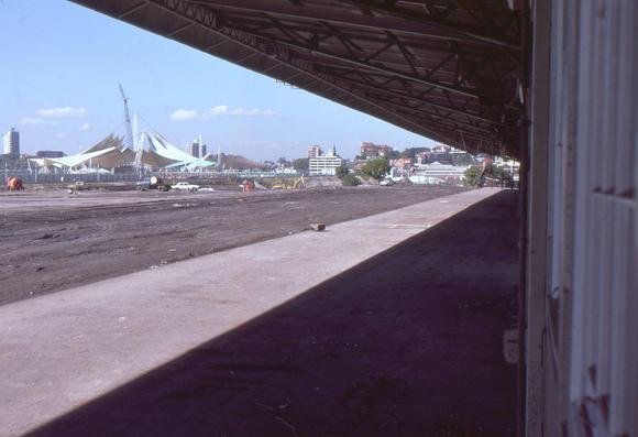 A view of a disused passenger railway platform taken from the passenger entrance gate.  Looking along the platform to our right, the wall of the station building is on the right, and the wide and completely deserted platform is to the left with the side nearest the building in shadow beneath the awning and the left part in bright sunlight.  All the tracks have been lifted and the land beyond the platform has been cleared.  A stone block has been placed on the platform edge, presumably an early part of the demolition process.  In the distance, beyond the former railway land, stands a structure made of fabric sails.