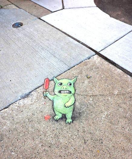 Streetart. On a gray sidewalk with four small holes in the ground, a small green monster (named Maurice) was drawn on the ground with chalk. The four holes have been integrated into the drawing and are now the eyes of the green friend. He is standing there looking scared with his mouth open and holding a red popsicle in his hand. A bee flies next to him. The ice cream is already dripping down. Title: 