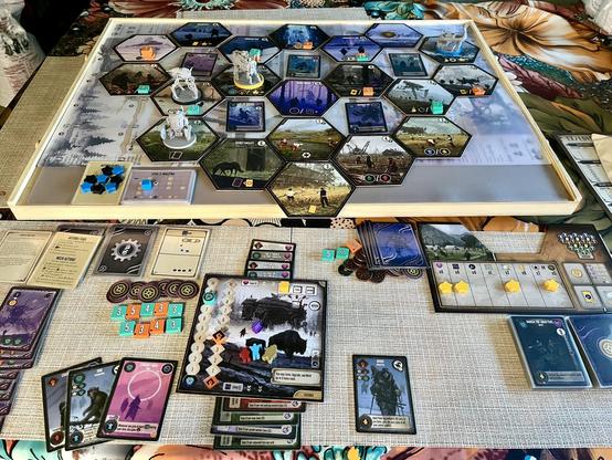 A strategic board game, Expeditions, is setup with a hexagonal map, game pieces, cards, tokens, and player boards arranged on a tabletop.